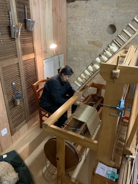 La tisserande au travail sur son métier à tisser en bois, devant le mur de pierre de l'atelier